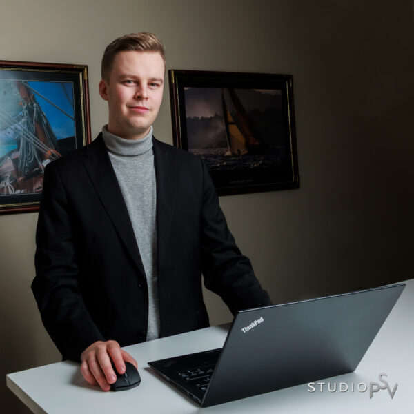 Nuori lakimies seisoo työpöydän takana ja hymyilee pienesti kameralle, pöydällä on avonainen lpääri