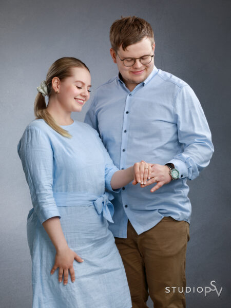 Nuori kihlapari katsoo kihlasormusta vaaleansinisissä asuissaan ja hymyilee