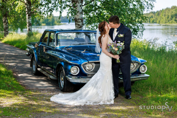 Hääpari suutelee toisiaan joen rannalla sinisen hääauton edessä ympärillään aurinkoinen ja kesäinen miljöö.