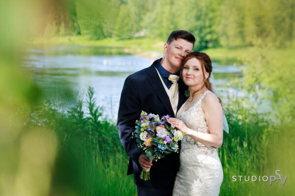 Hääpari nojaa toisiinsa ja poseeraa rannalla ympärillään paljon vihreää.