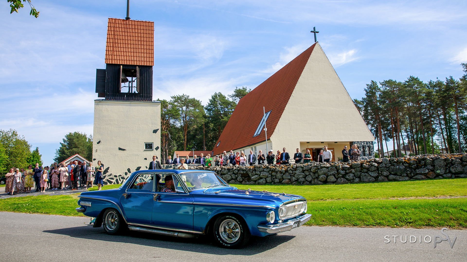 Sininen auto, jossa istuu hääpari, etualalla ja kirkko hääväen kanssa taka-alalla
