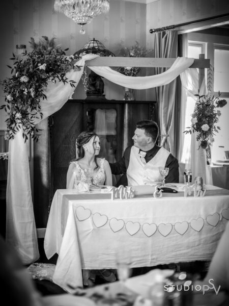 Hääpari istuu morsiuspöydässä, jonka ylle on rakennettu kukista ja harsoista portti. Pari katsoo kauniisti toisiaan.