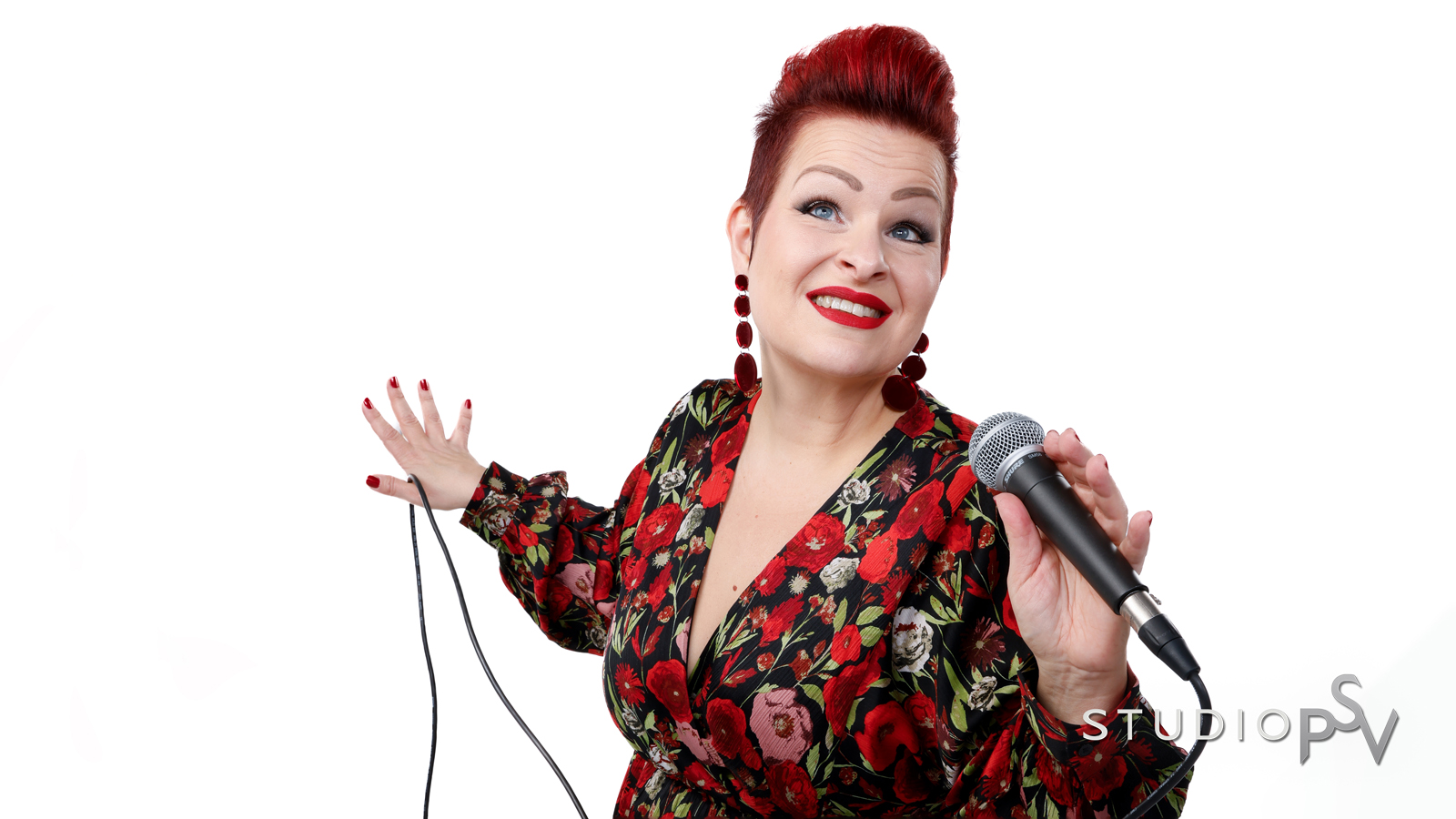 Punahiuksinen nainen punaisessa kukkamekossa mikrofoni kädessä valkoista taustaa vasten